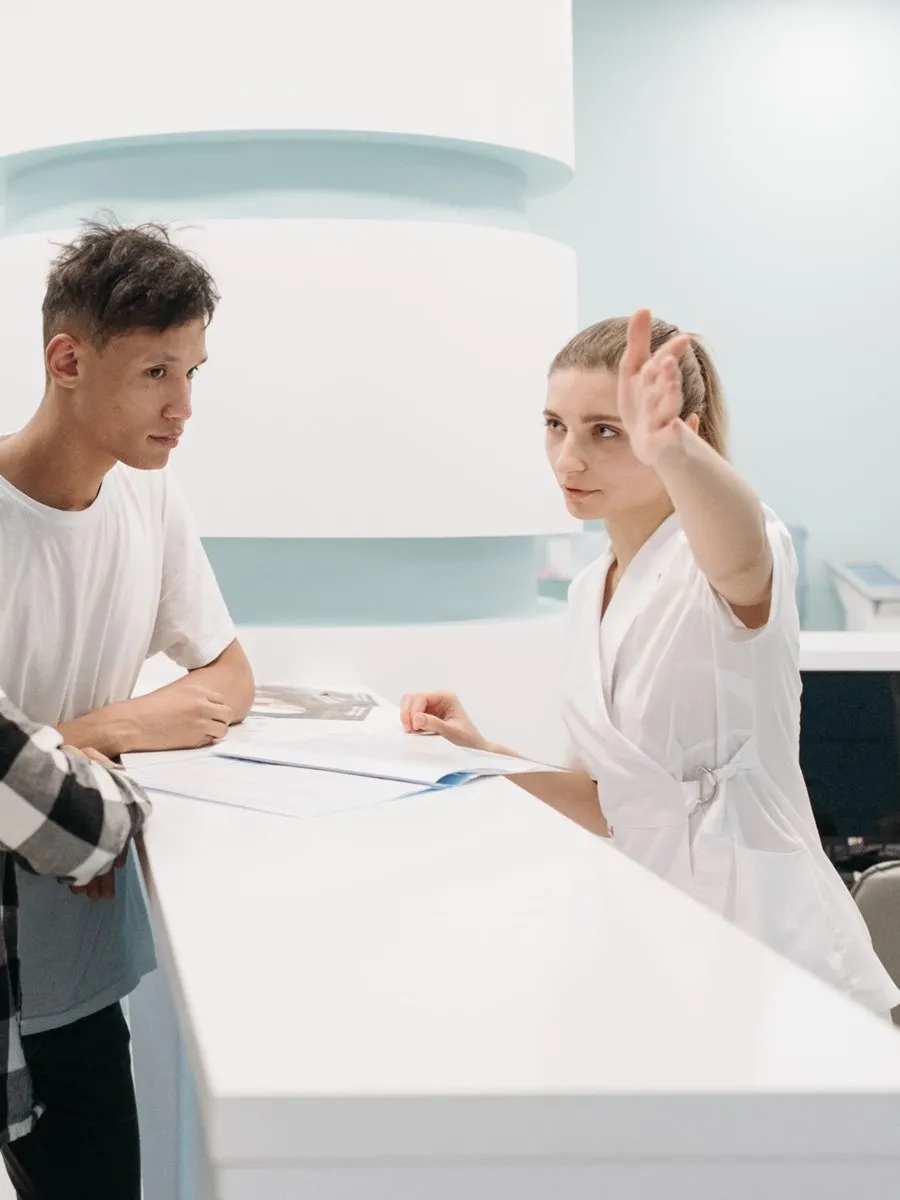A receptionist in a white uniform at a clinic front desk, pointing a patient the right way