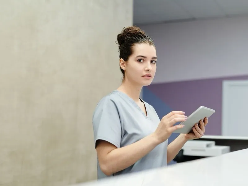A clerk in a light blue uniform holding a tablet at a hospital reception desk